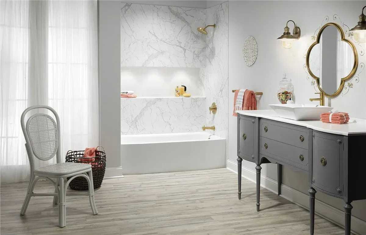Bright bathroom with a freestanding tub, gray vanity, gold mirror, and wicker chair by a curtained window.