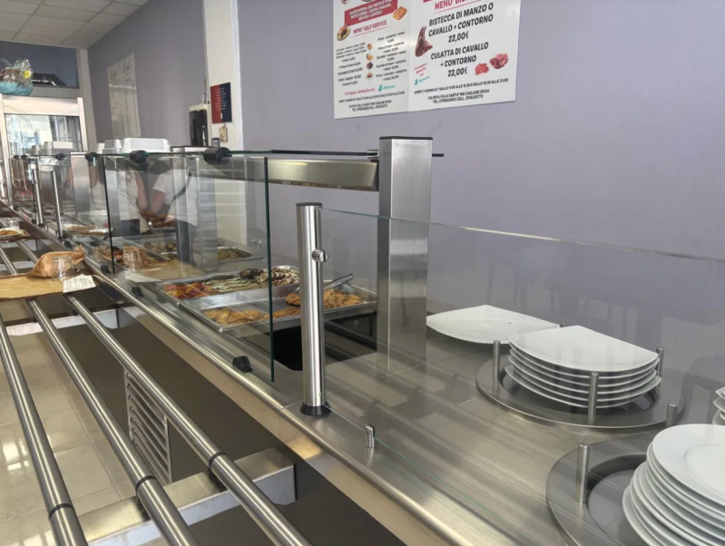 Uomo al lavoro con un’affettatrice in una salumeria, con tagli di carne appesi alle sue spalle.