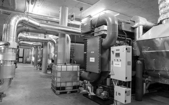 Industrial mechanical room with large silver ducts, pipes, and utility equipment along a concrete floor.