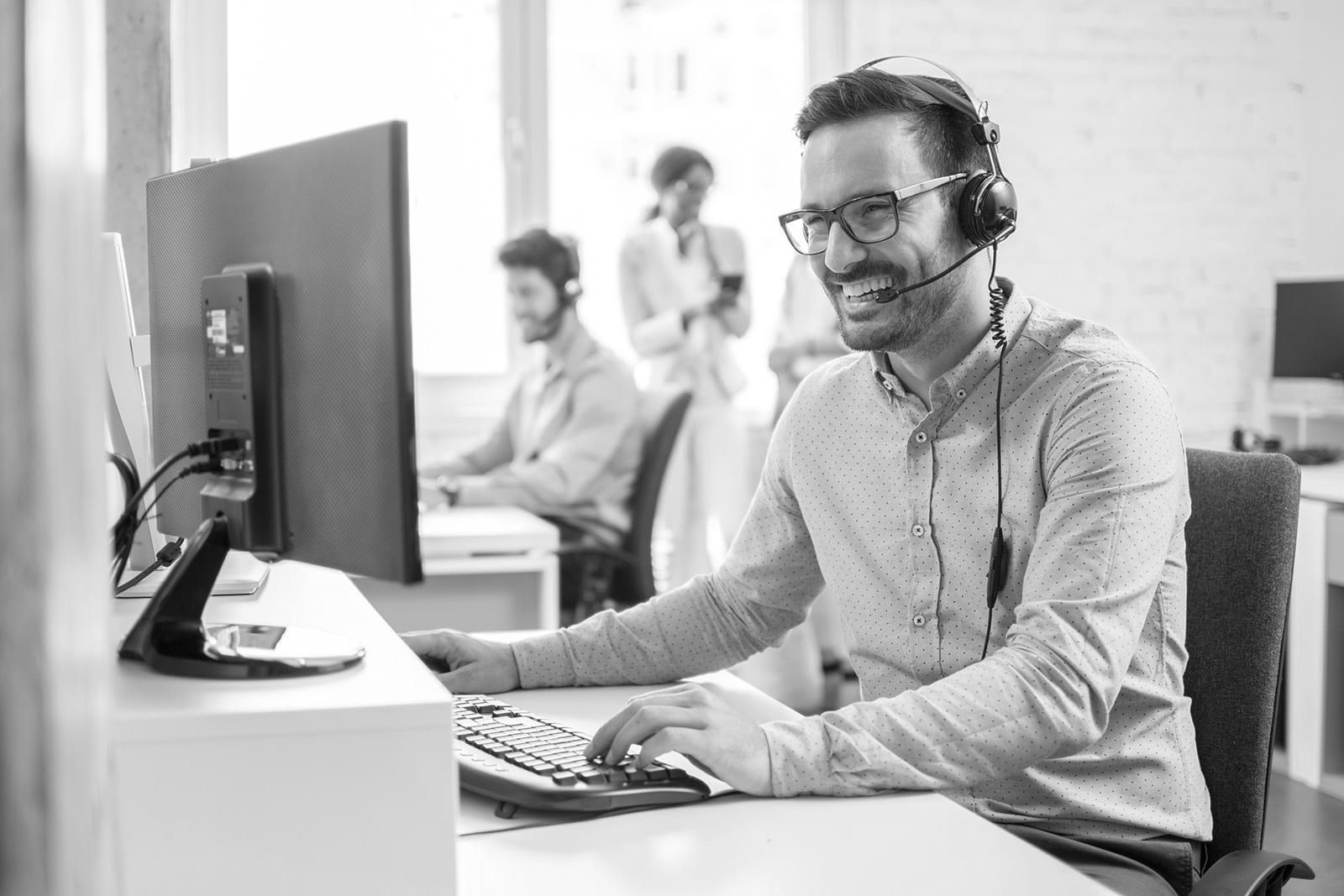 Man smiling at a computer in a bright office with coworkers blurred in the background