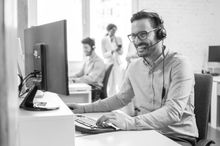 Man smiling at a computer in a bright office with coworkers blurred in the background
