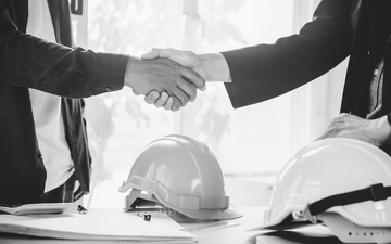 Two people in business suits shaking hands across a table with hard hats, suggesting a construction deal