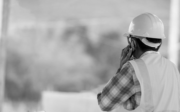 Worker in hard hat and safety vest using a phone at a construction site, with dust in the background