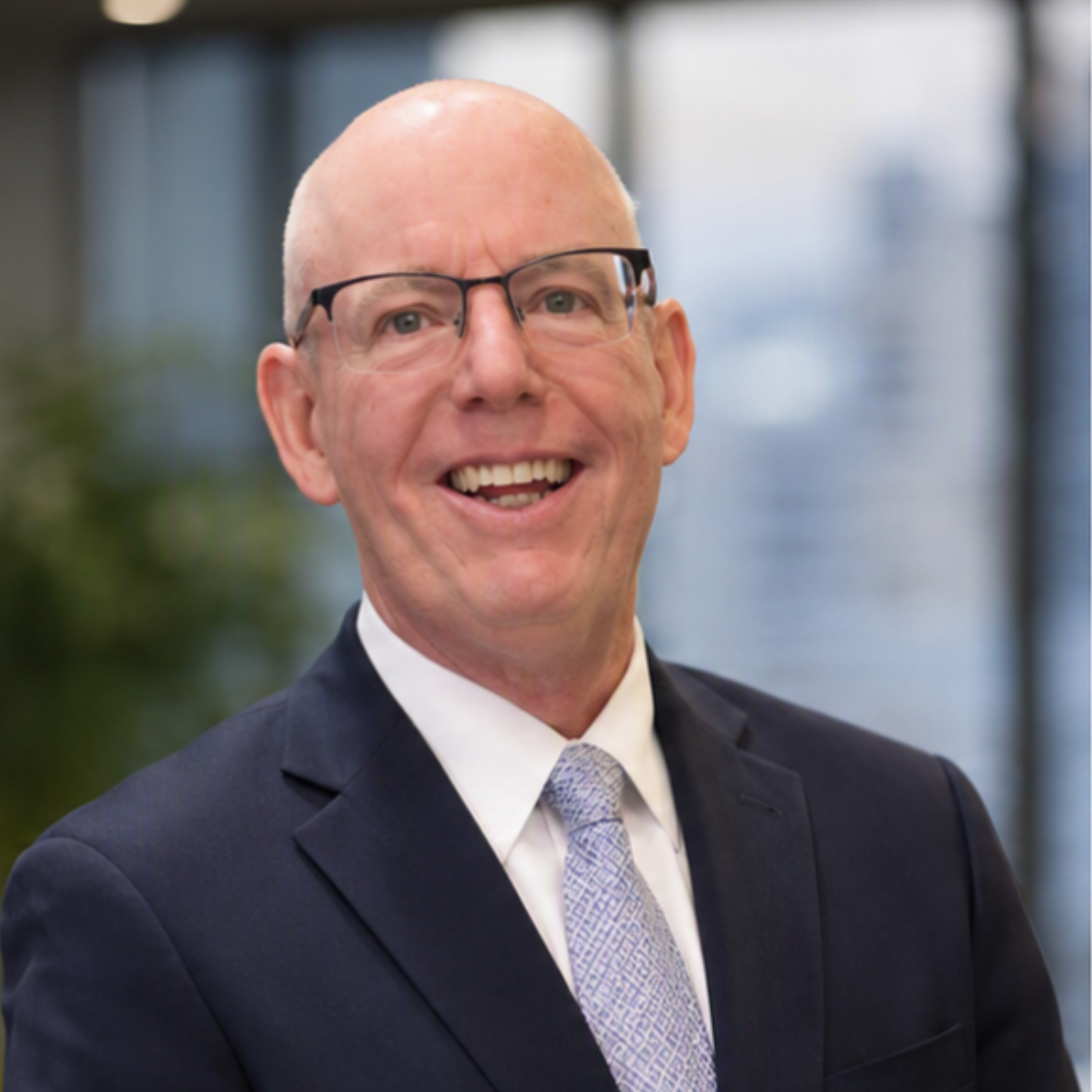 Smiling bald man in glasses wearing a navy suit and tie against a blurred office background