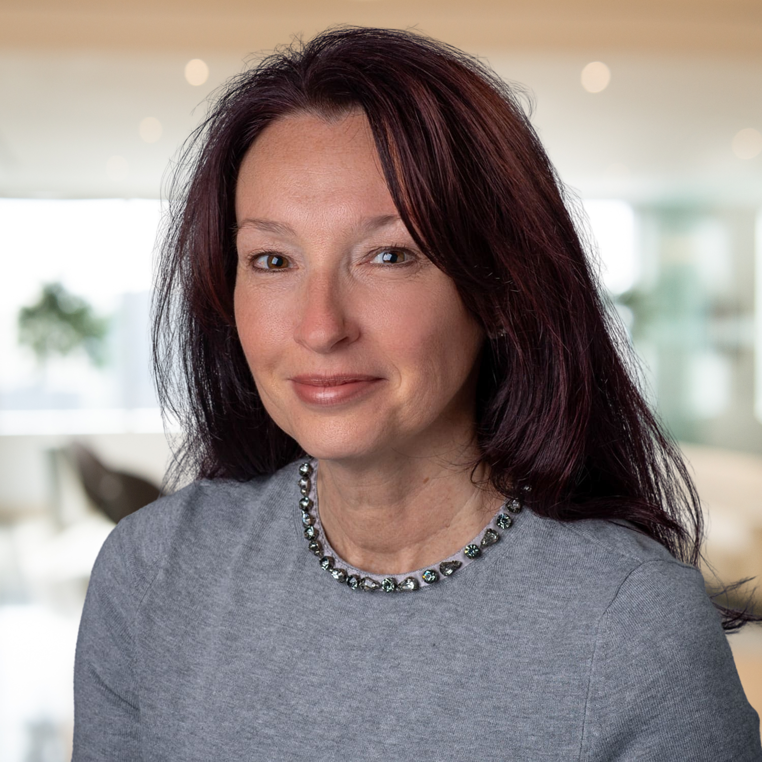 Portrait of a woman with dark hair in a gray top, wearing a beaded necklace, in a softly lit indoor setting