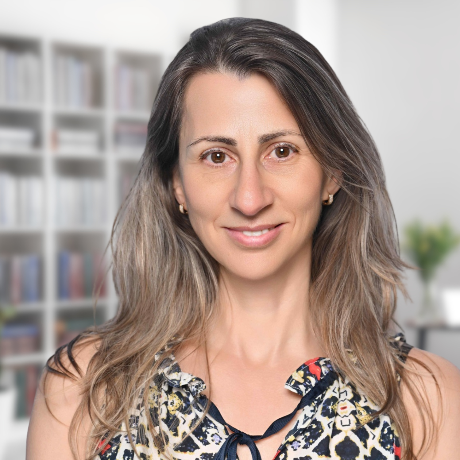 Portrait of a smiling woman with long hair in a patterned blouse, indoors with shelves blurred behind her