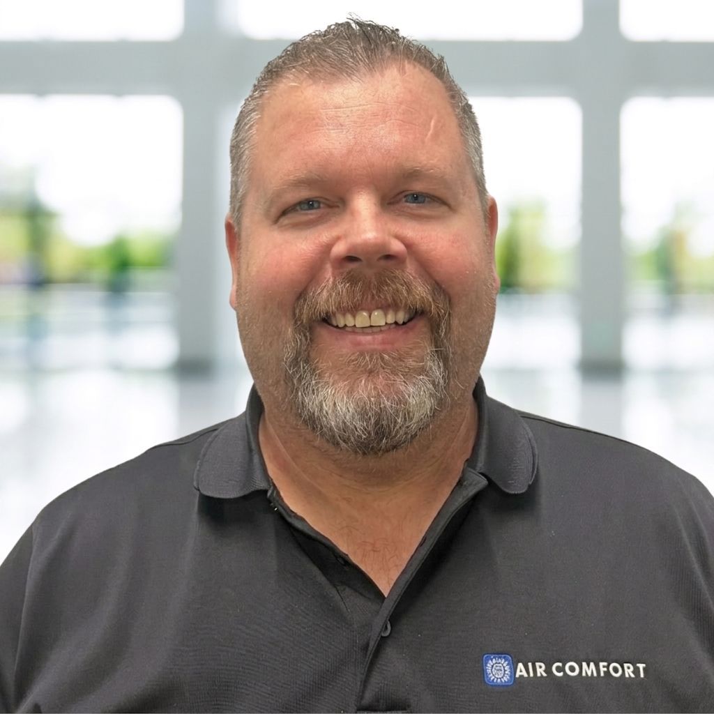 Smiling man in a dark polo shirt with an Air Comfort logo, standing in front of a bright blurred office background