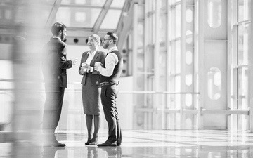 Three businesspeople talking in a bright glass atrium, with one gesturing while the others listen.