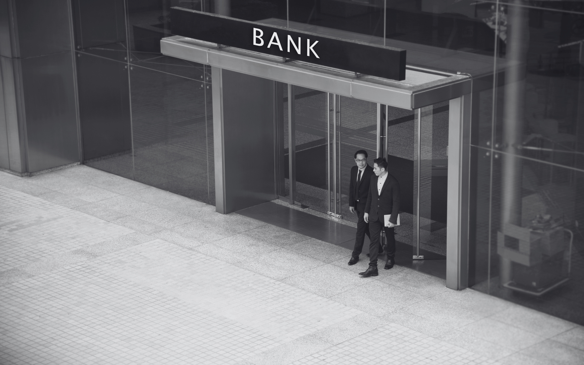 Two people stand at a bank entrance under a BANK sign.