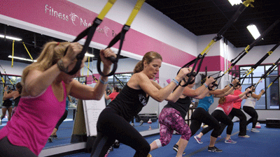 A group of people in an exercise studio performing TRX suspension training with straps attached to the ceiling.