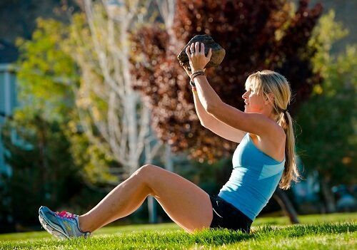 A person in a blue tank top and black shorts sits on a grassy lawn performing a sit-up while holding a heavy rock.