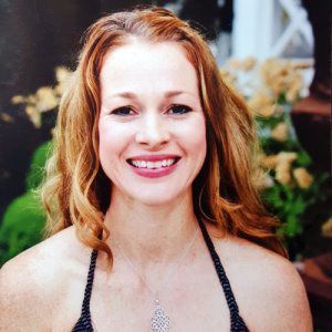 A smiling person with wavy red hair wearing a dark halter-neck top and a silver pendant necklace against a garden background.