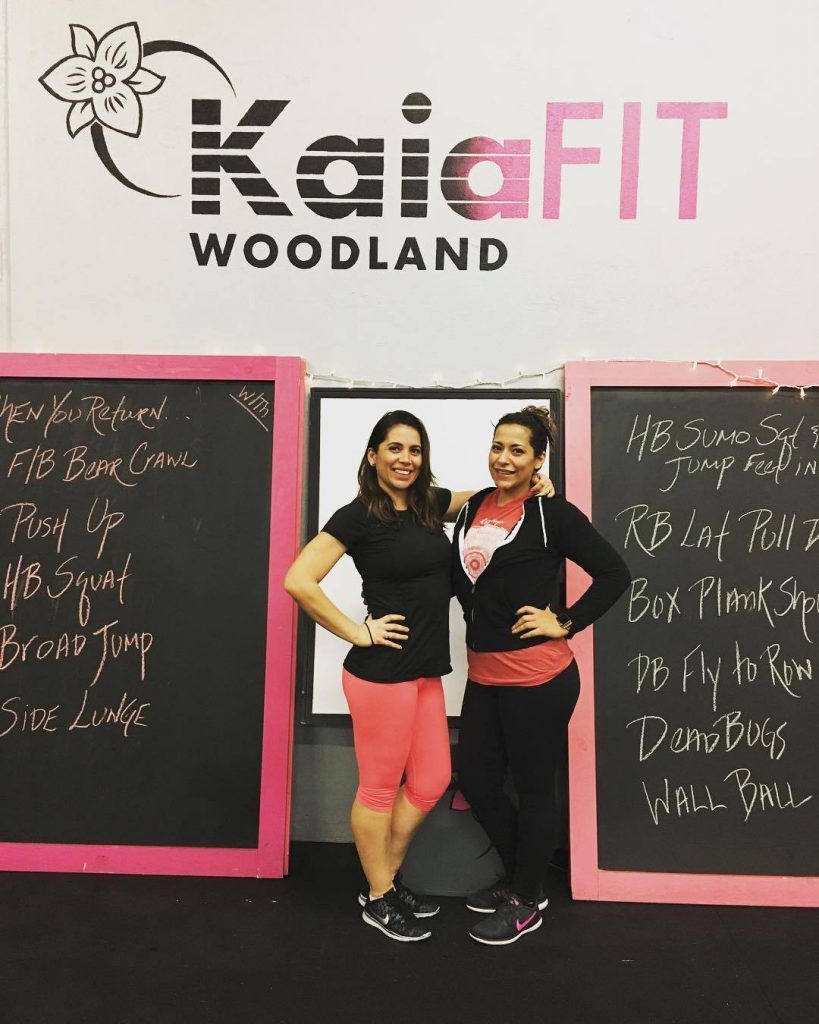 Two women smiling and posing in front of a KaiaFIT Woodland sign flanked by two chalkboards listing exercise routines.