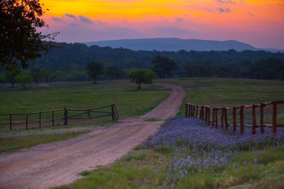 A dirt road going through a field with a fence and a sunset in the background.