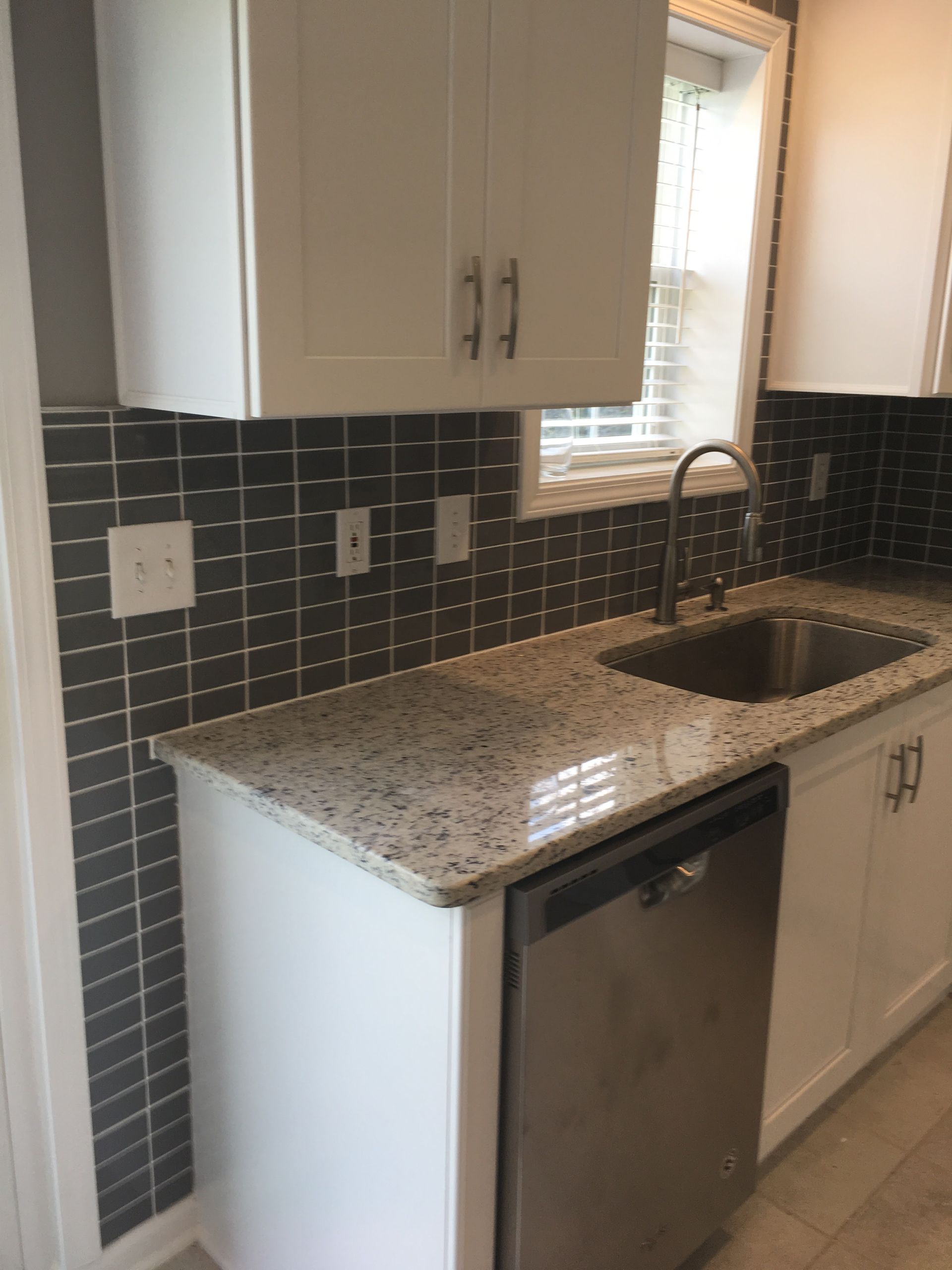Kitchen with white cabinets, speckled countertop, stainless steel sink, and dark gray tile backsplash.