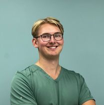 Tannlege Christian med briller og skjegg, iført blå skjorte og medisinsk maske, smiler til kameraet.
