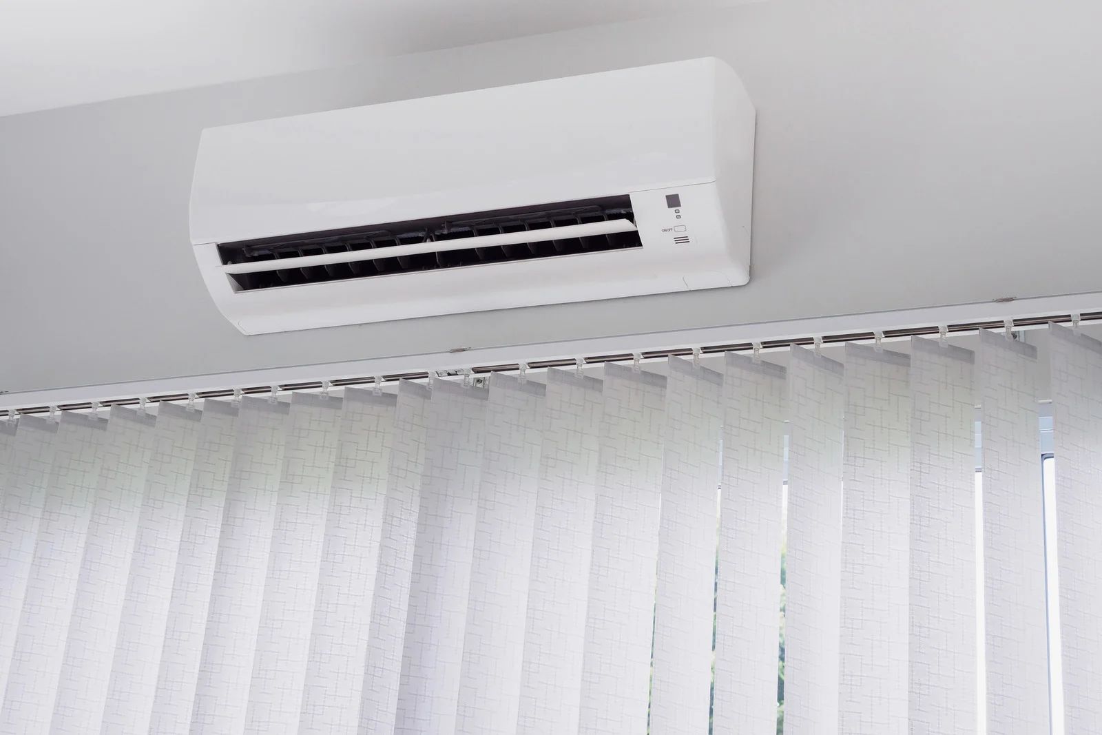 Wall-mounted air conditioner above white vertical blinds in a bright room