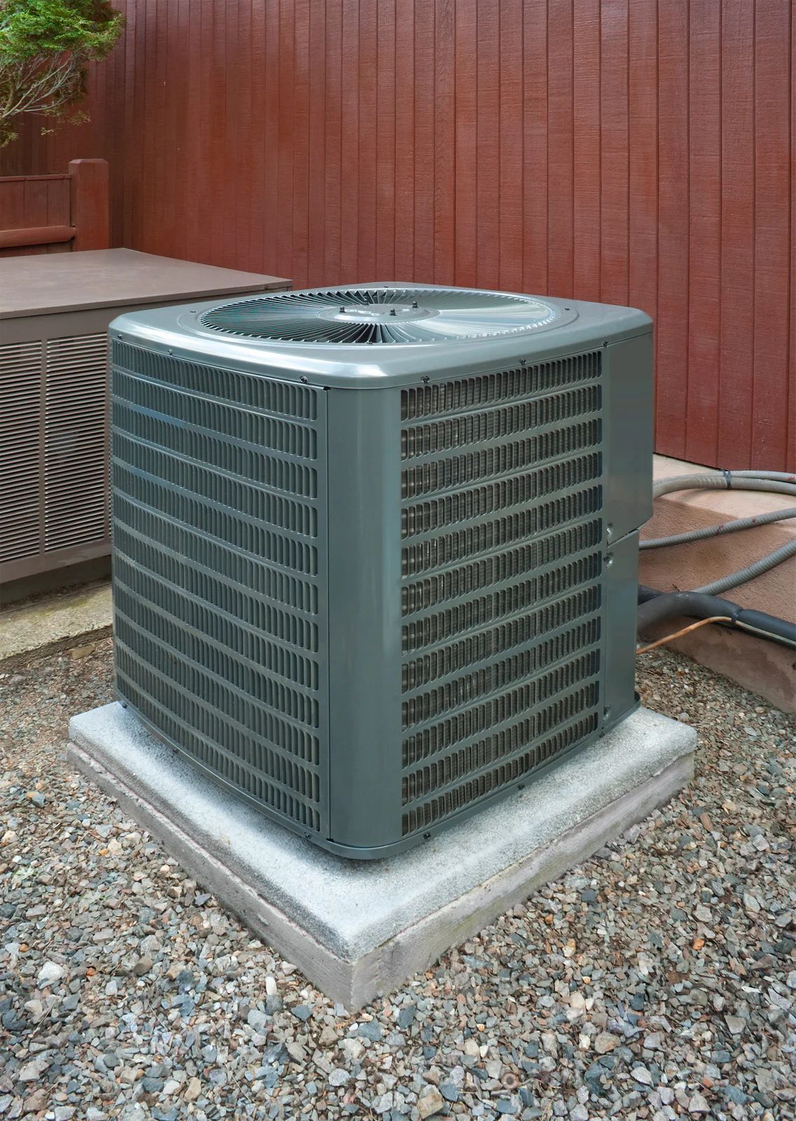Outdoor air conditioning unit beside a wooden wall on a gravel pad.
