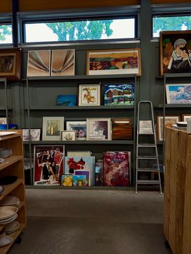 In een winkel staan planken vol ingelijste kunstwerken; een ladder leunt tegen de muur.