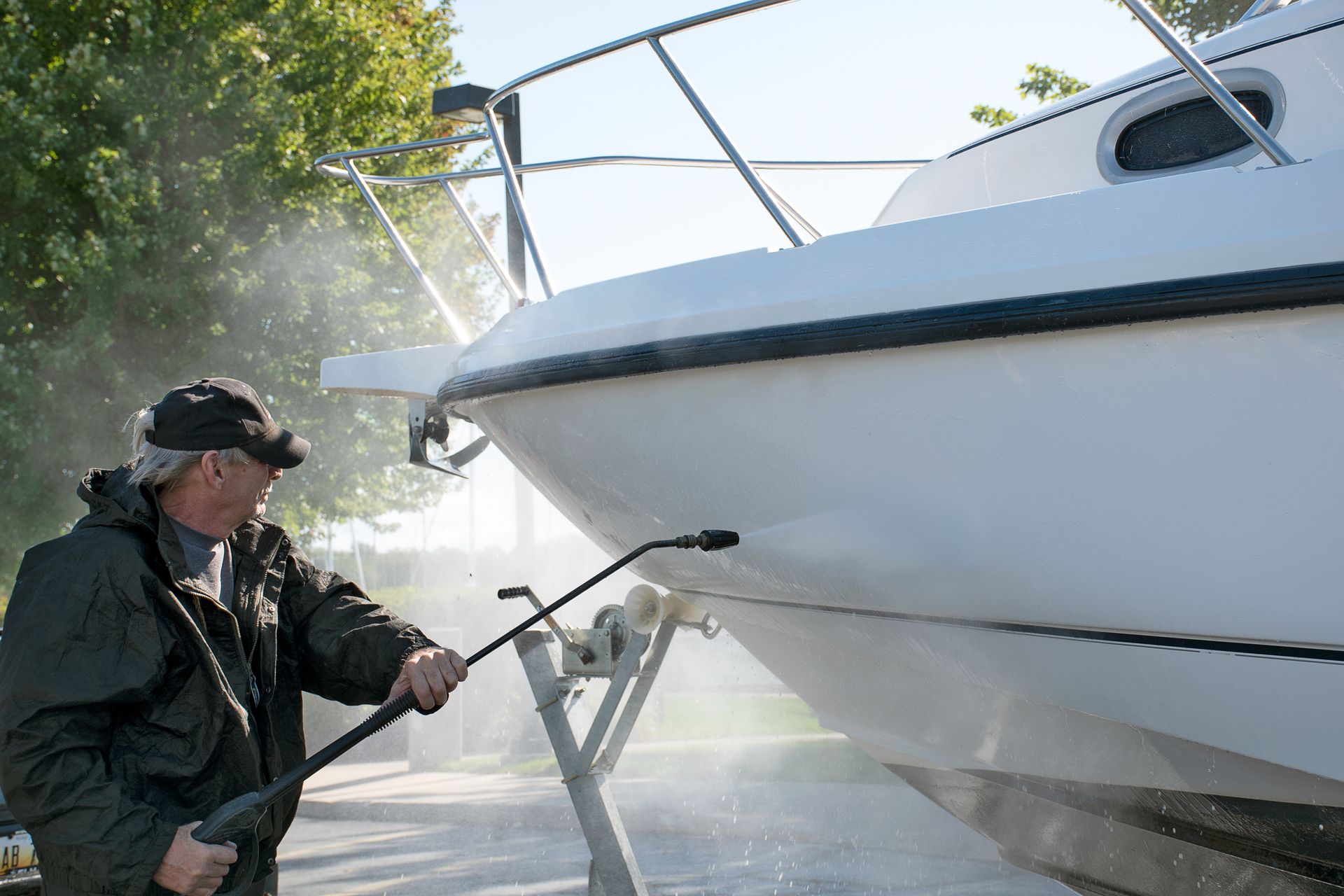 Man in a black jacket power washing a white boat on a trailer outdoors.