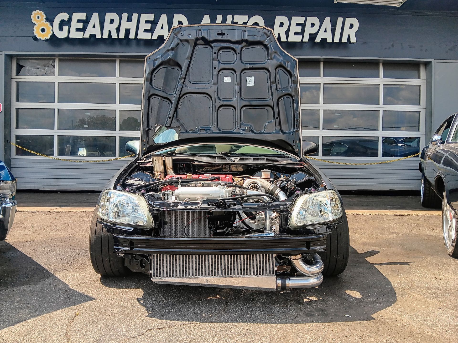 Close-up of a car's exhaust pipe emitting white smoke. Black car with hood open, showing engine modifications, in front of