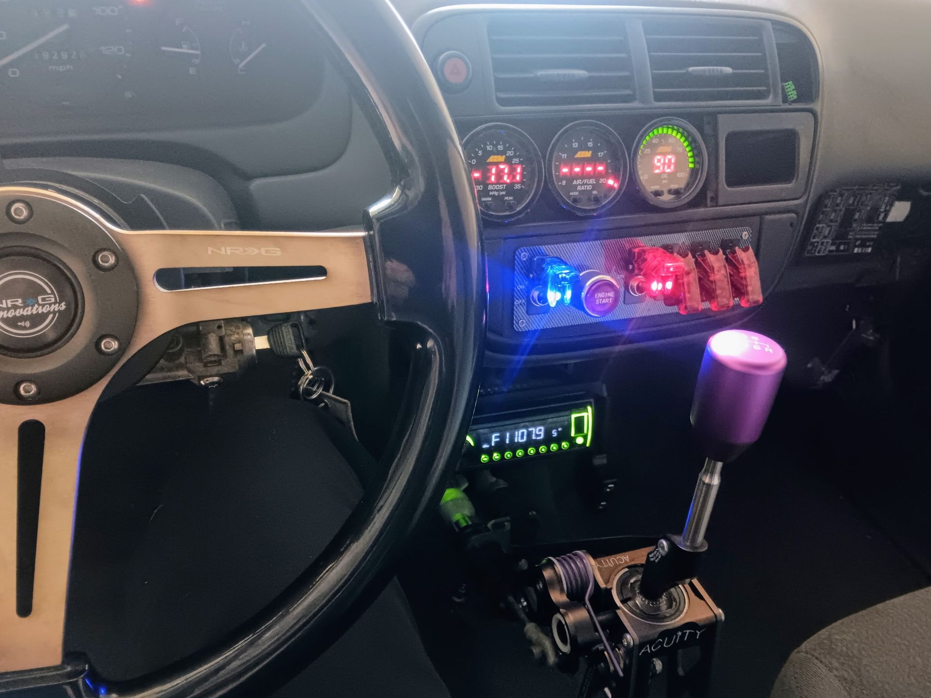 Hand turning the dial on a car radio, close-up shot of the dashboard. Car interior: steering wheel, gauges, switches, gear shift. Purple and blue accents.