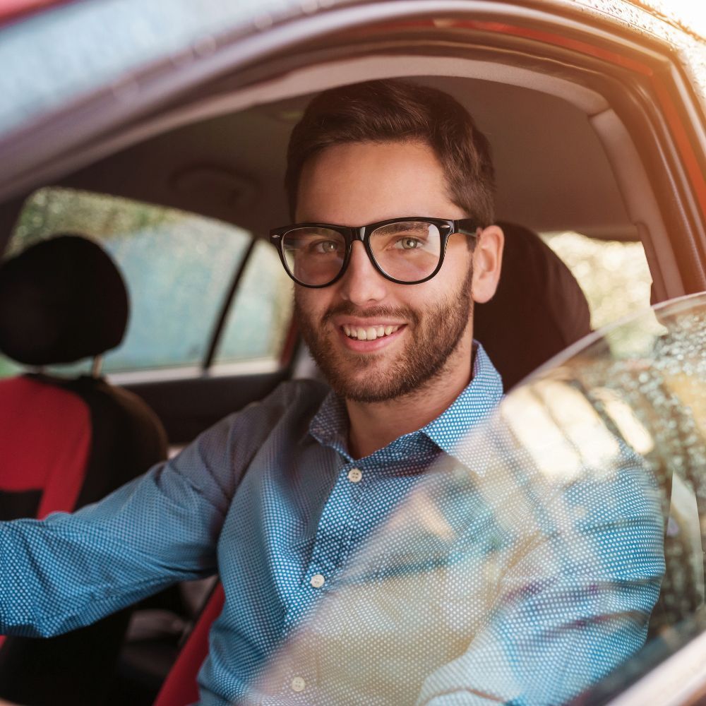 Man in glasses smiling, sitting in a red car, looking at the camera.