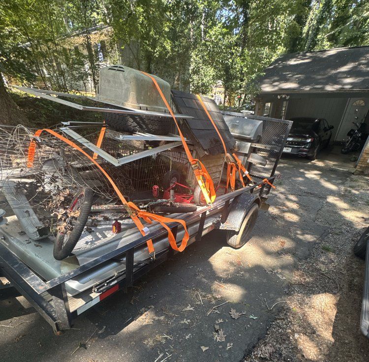 Flatbed trailer loaded with strapped metal parts in a driveway beside a garage, under trees.