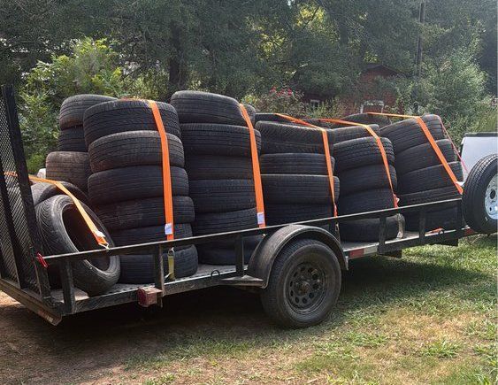 Trailer loaded with stacks of tires strapped down with orange tie-downs outdoors.