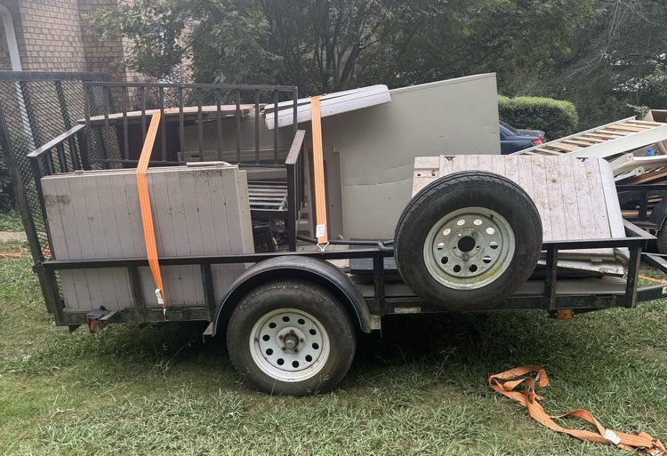 Small trailer loaded with scrap metal and furniture in a yard, secured with orange straps.