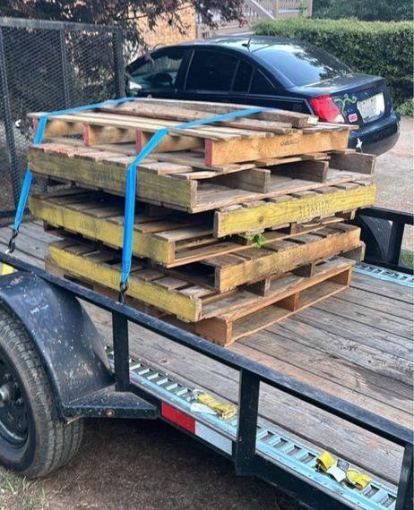 Stacked wooden pallets strapped on a utility trailer, with a black car parked behind.