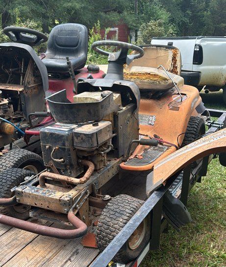 Rusty orange riding mower with a black seat and damaged engine, parked outdoors in a grassy yard.