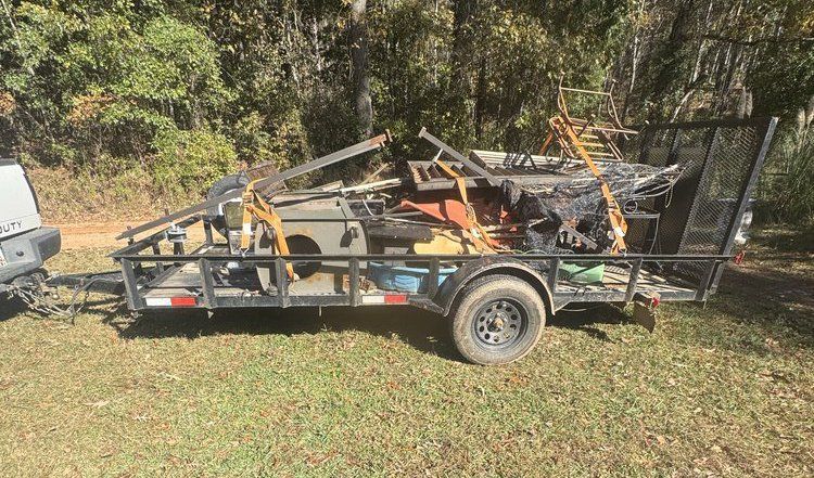 Flatbed trailer loaded with scrap metal and debris, parked on a grassy area near trees.