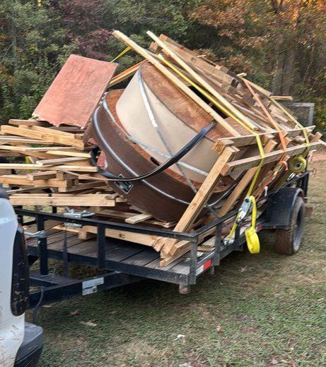 Overloaded trailer carrying lumber and debris, with a large tipped-over wooden tub outdoors.