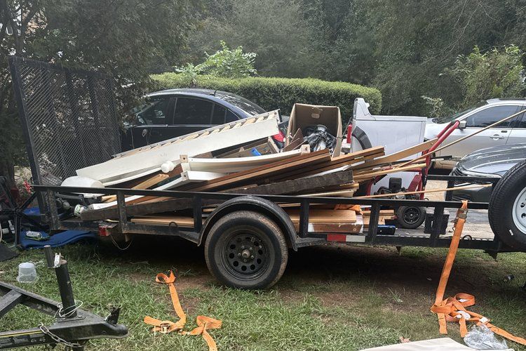 Trailer piled with scrap wood and debris parked on grass beside two cars and trees