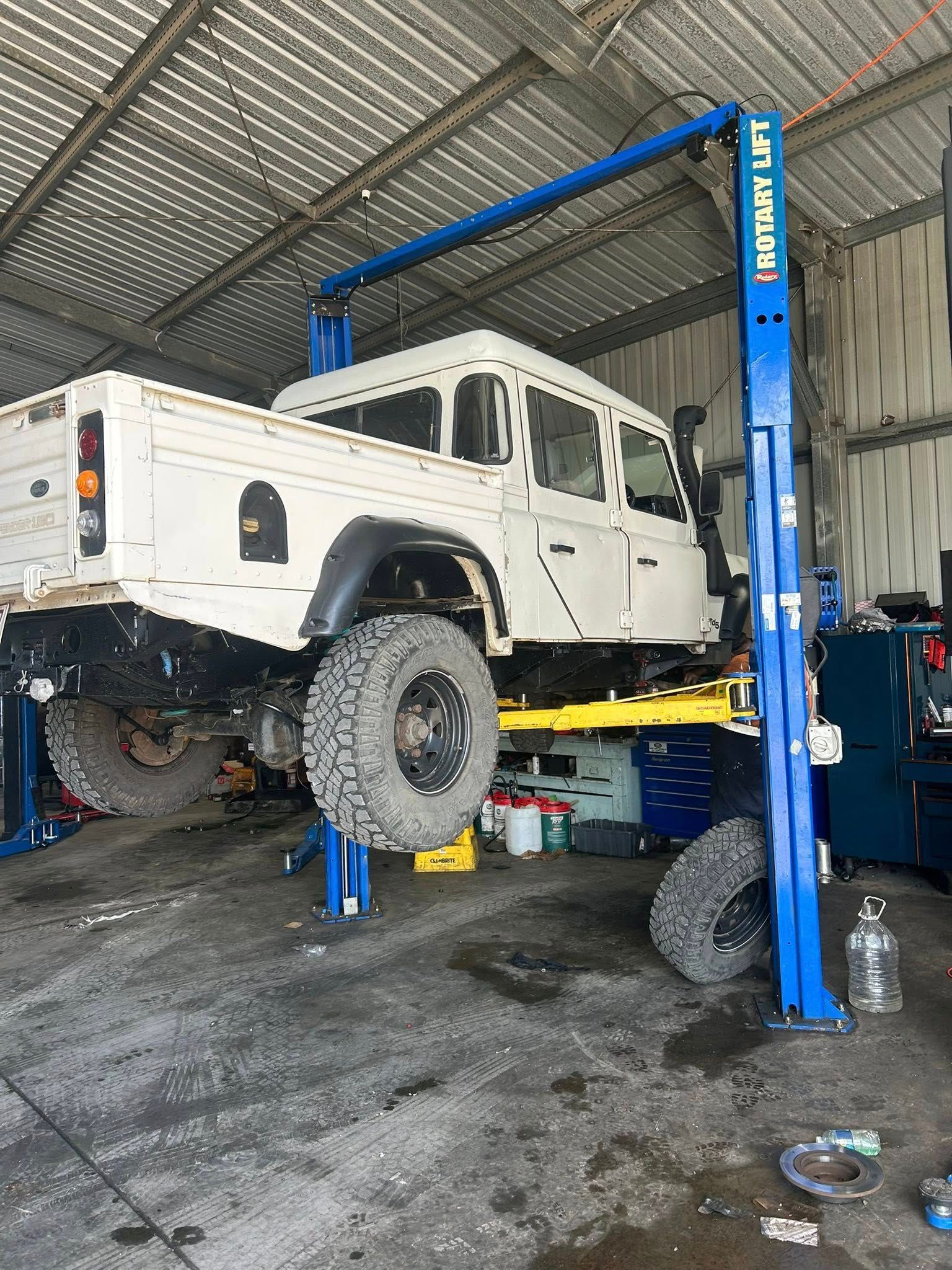 White truck on a blue auto lift in a garage. Large off-road tires, metal roof.
