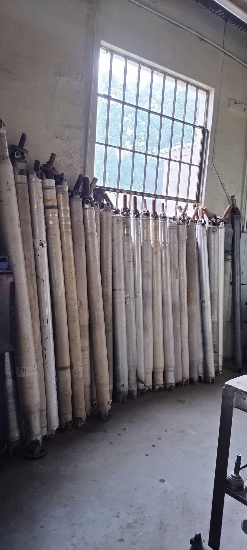 A row of tall, cylindrical industrial drive shafts lined up against a wall in a workshop beneath a grid-paned window.