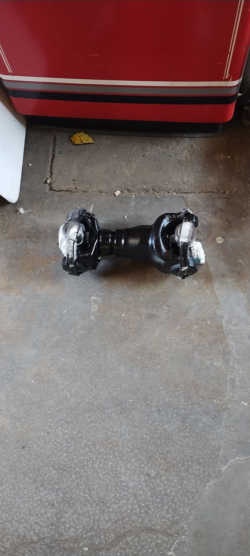 A black metal driveshaft with universal joints at both ends resting on a grey concrete floor.