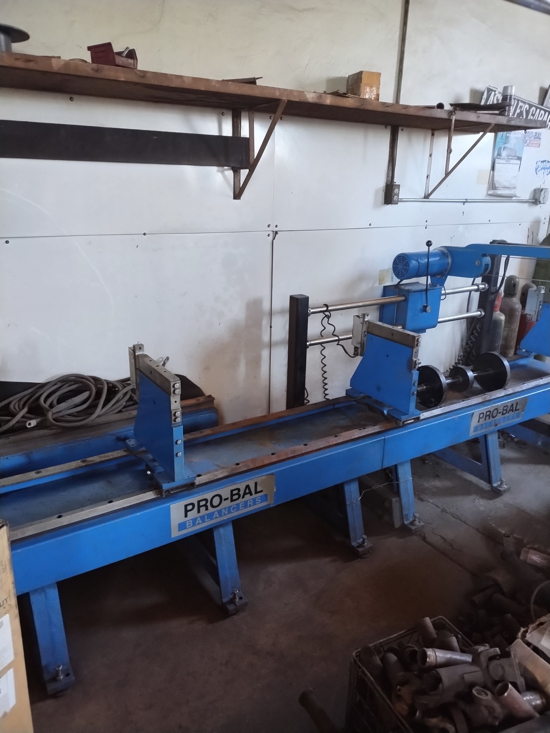 A blue Pro-Bal industrial driveshaft balancing machine sits in a workshop with shelves in the background.