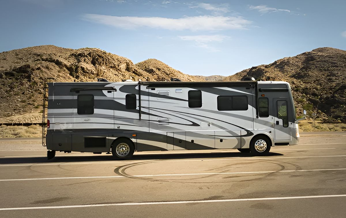 A large, gray and white recreational vehicle parked on an asphalt lot in front of rocky desert hills.