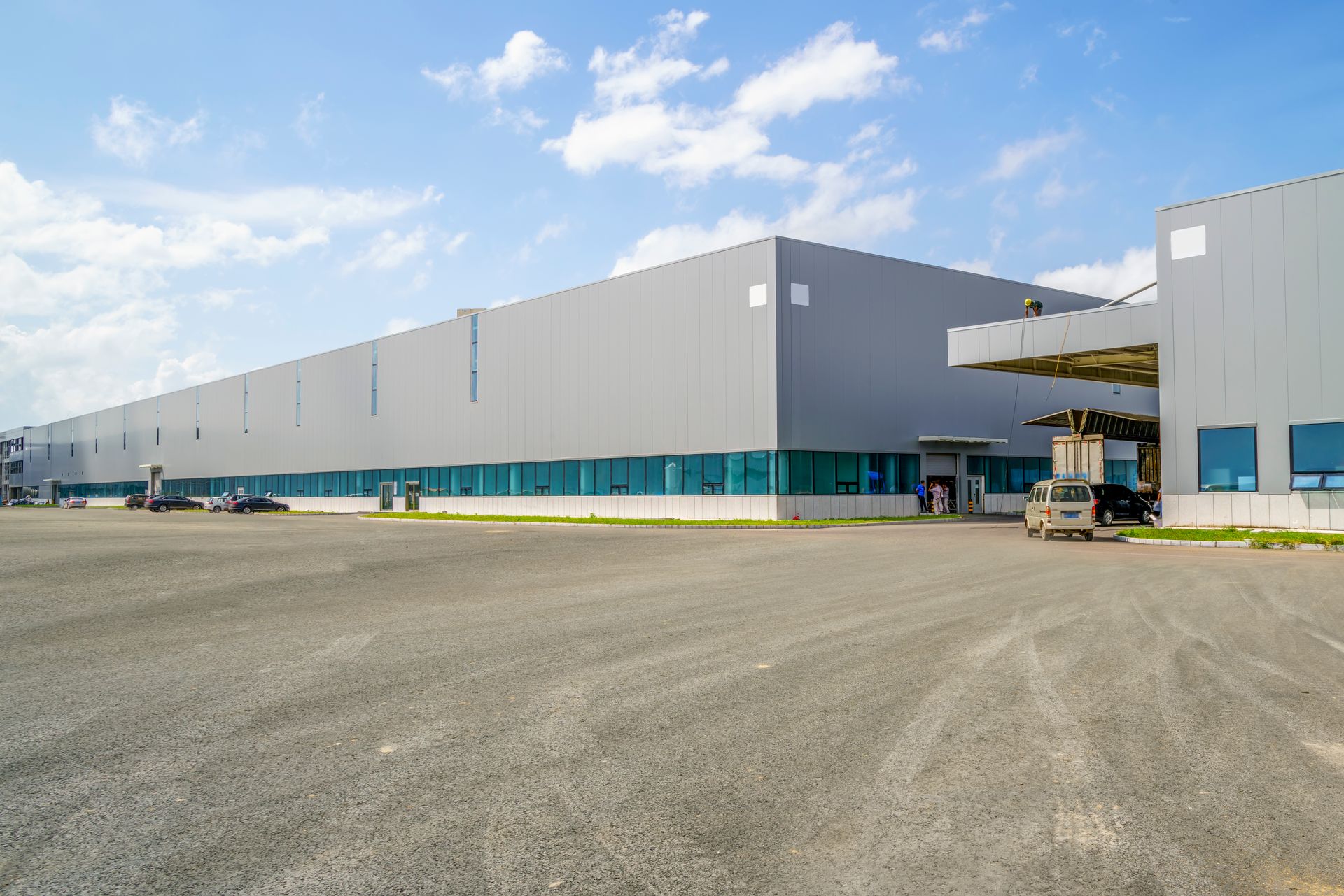 A long, gray, modern industrial warehouse building with a paved parking lot under a blue sky.