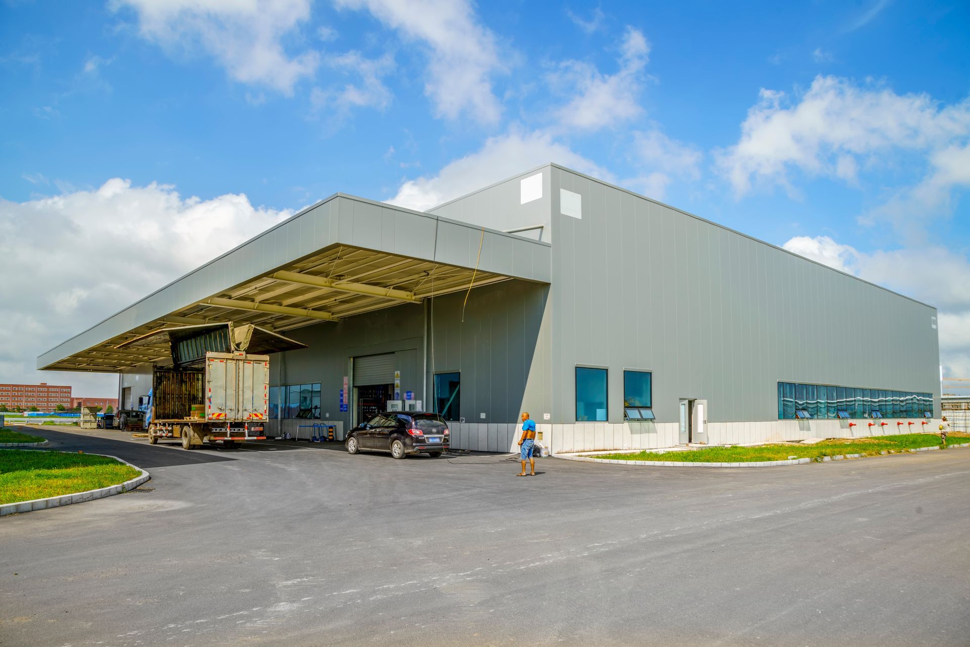 A modern gray warehouse exterior with a large loading bay, a truck being loaded, and a parked car under a sunny blue sky.