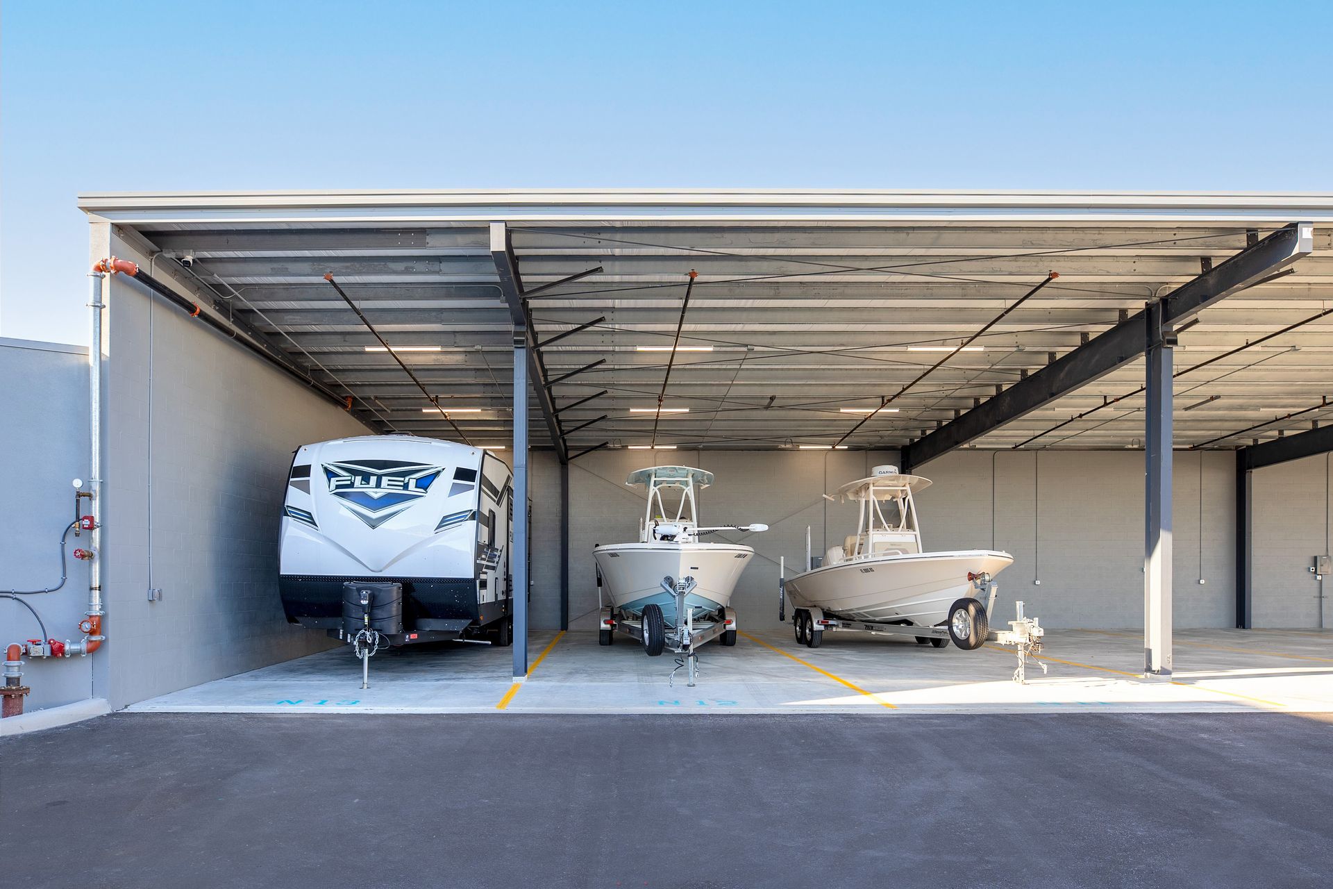 An open-air storage facility shelters a camper and two boats under a metal roof; the setting is outdoors.