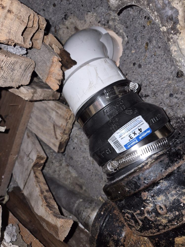 Broken PVC pipe fitting in a wall opening, with a black connector and white elbow joint among rubble.