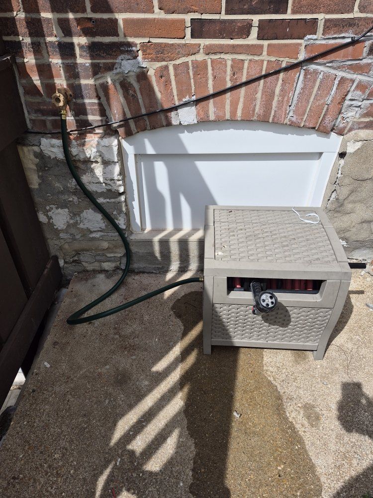 Beige outdoor utility box on a concrete patio beside a brick wall, with a hose and brick shadows nearby