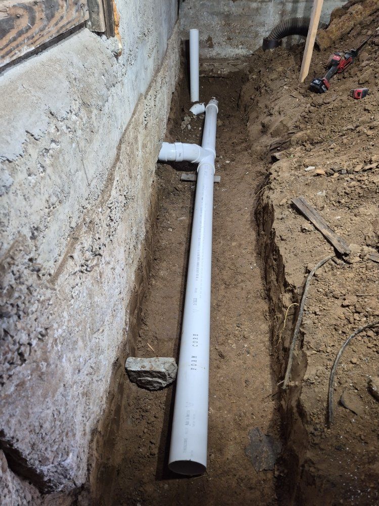 White PVC pipes laid in a dirt trench beside a foundation wall during plumbing installation