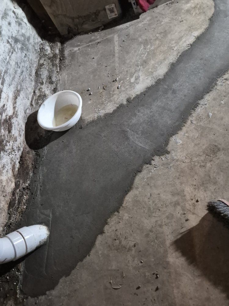 White bowl on a rough concrete floor beside a wall, with a pipe at the lower left.