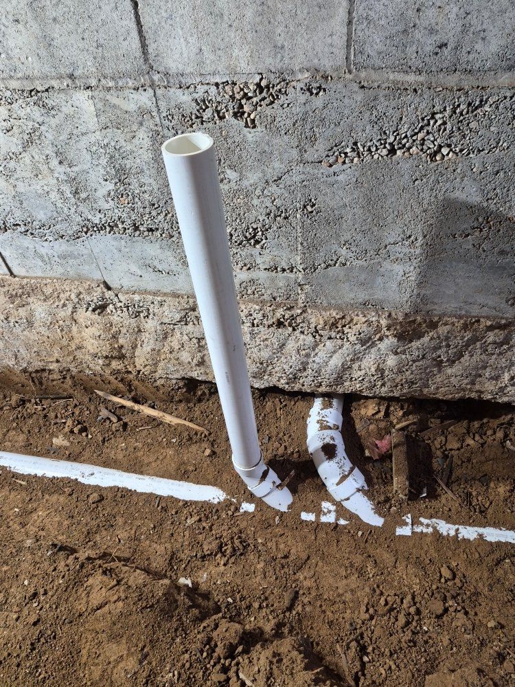 White PVC pipe stub and broken pipe fittings emerging from soil beside a concrete wall.
