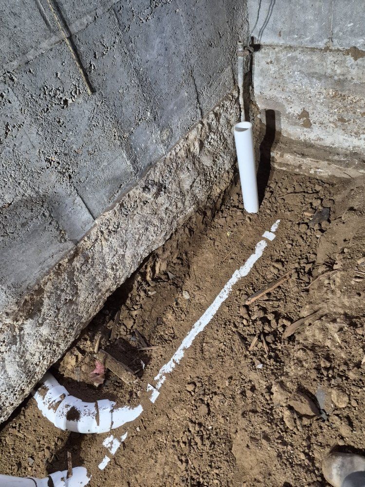 Trench beside a stone wall with exposed white PVC pipe and muddy soil around a drain opening
