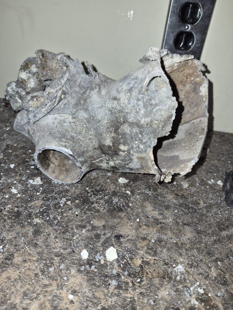 Cracked, corroded pipe fitting on a dirty floor beside a metal support bracket